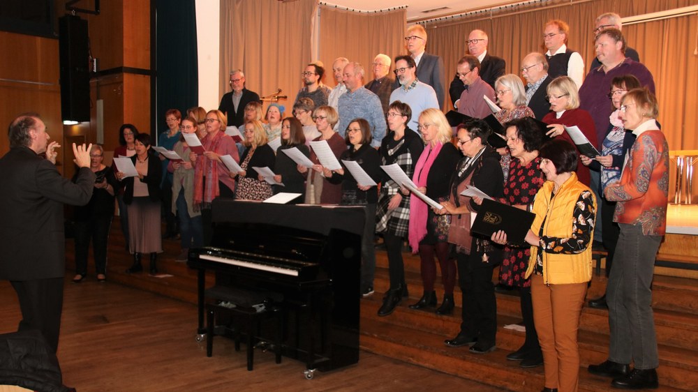 Die SVM-Chöre singen bei der Weihnachtsfeier mit Notenblättern auf den Stufen der Bürgerhausbühne, links der Dirigent, in der Mitte ein Klavier