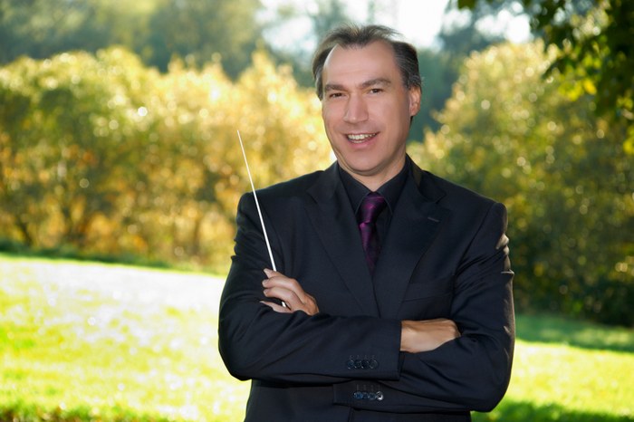 Chorleiter Werner Utmelleki im dunklen Anzug mit Taktstock, im Freien vor herbstlichen Bäumen