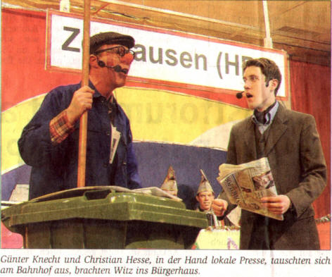 Günter Knecht und Christian Hesse mit Zeitungen in der Hand beim Sketch am Bahnhofsschild Zellhausen, Zeitungsausschnitt mit Bildunterschrift