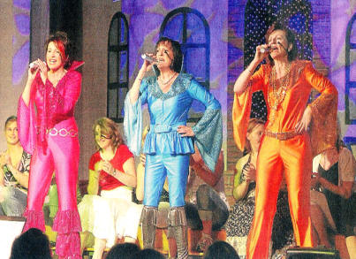 Drei Sängerinnen in pinkfarbenem, blauem und orangefarbenem Glitzerkostüm im ABBA-Stil singen mit Mikrofonen auf der Bühne