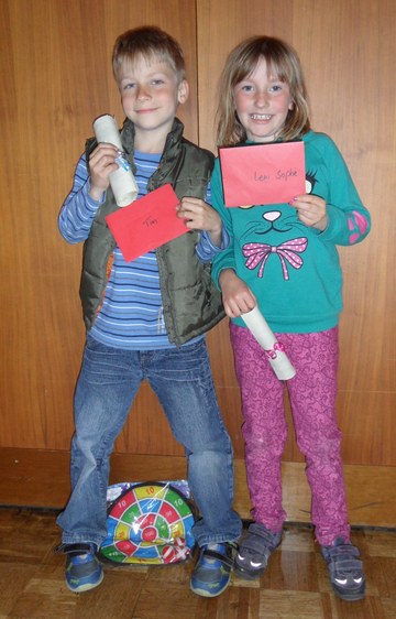Ein Junge und ein Mädchen aus dem Kinderchor mit Urkundenrollen und roten Umschlägen, auf denen die Namen Tim und Leni Sophie stehen, davor ein Dartspiel als Geschenk