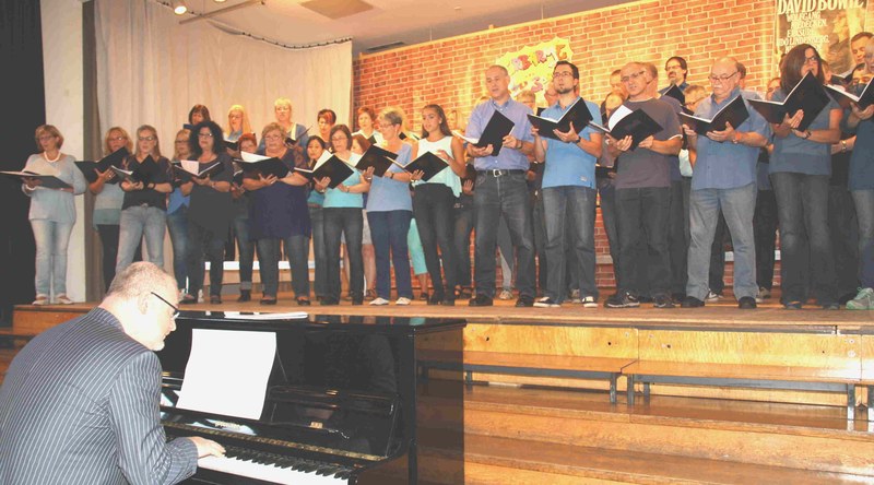 Der Chor in Blautönen singt mit schwarzen Notenmappen auf der Bühne vor einer Ziegelwand-Kulisse, vorne begleitet ein Pianist am Klavier