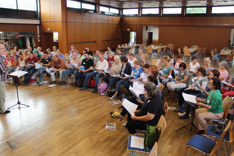 Die Workshop-Teilnehmer sitzen mit Noten im großen Halbkreis im Saal, links leitet der Dozent die Probe