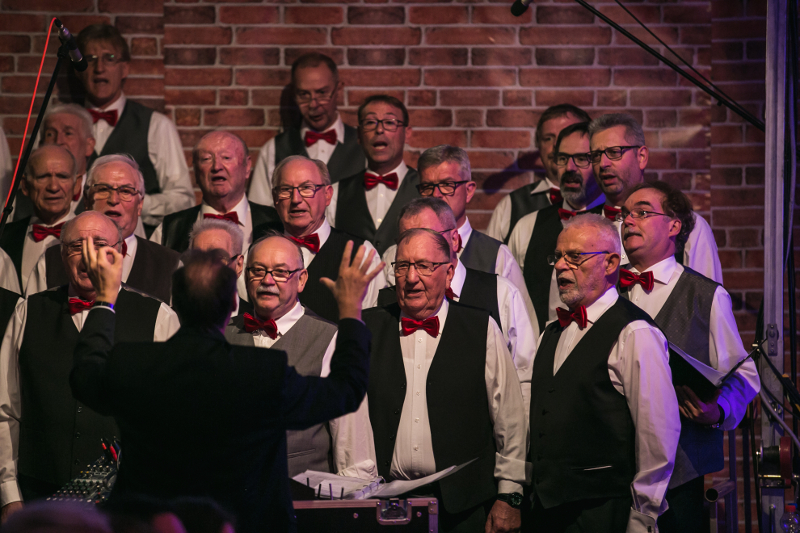Der Männerchor in weißen Hemden, dunklen Westen und roten Fliegen singt vor einer Backsteinkulisse, davor dirigiert der Chorleiter mit erhobenen Armen