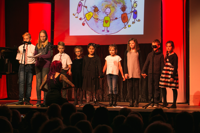 Der Kinderchor an Mikrofonen auf der rot beleuchteten Bühne, dahinter die Projektion einer gemalten Weltkugel mit Kindern, davor kniet die Chorleiterin