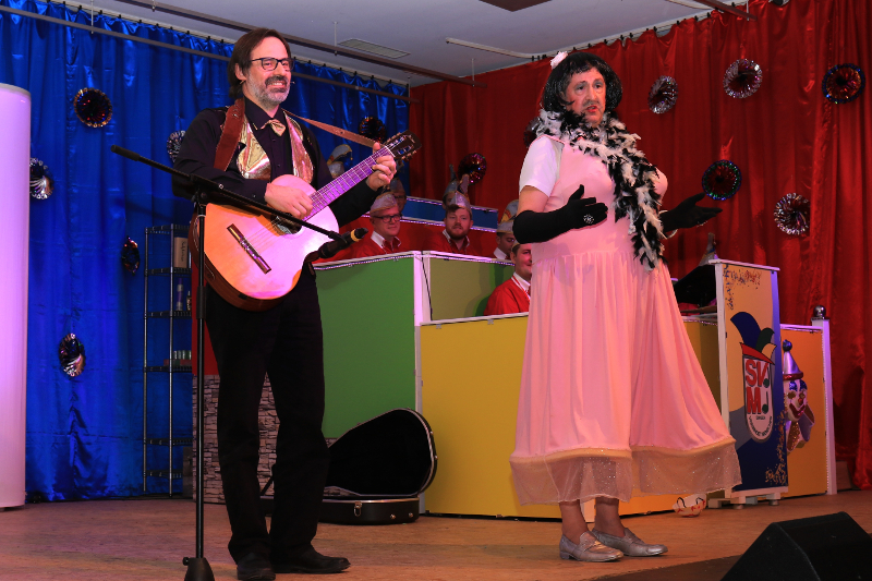 Zwei Büttenasse auf der Bühne: einer mit Gitarre am Mikrofon, der andere im rosa Kleid mit Federboa und schwarzen Handschuhen, dahinter der Elferrat