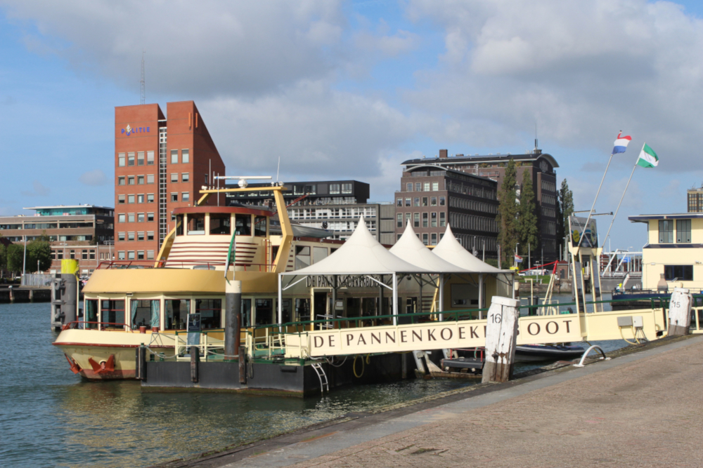 Das Pannenkoekenboot mit weißen Zeltdächern am Kai im Rotterdamer Hafen, dahinter moderne Bürogebäude und die niederländische Flagge