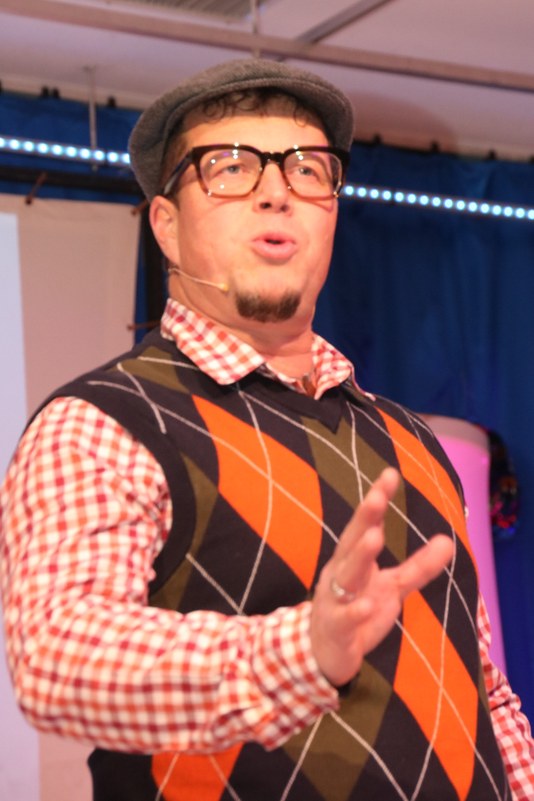 Christoph Gast in seiner Rolle als „Schambes“ mit Schiebermütze, Brille und Pullunder beim Vortrag