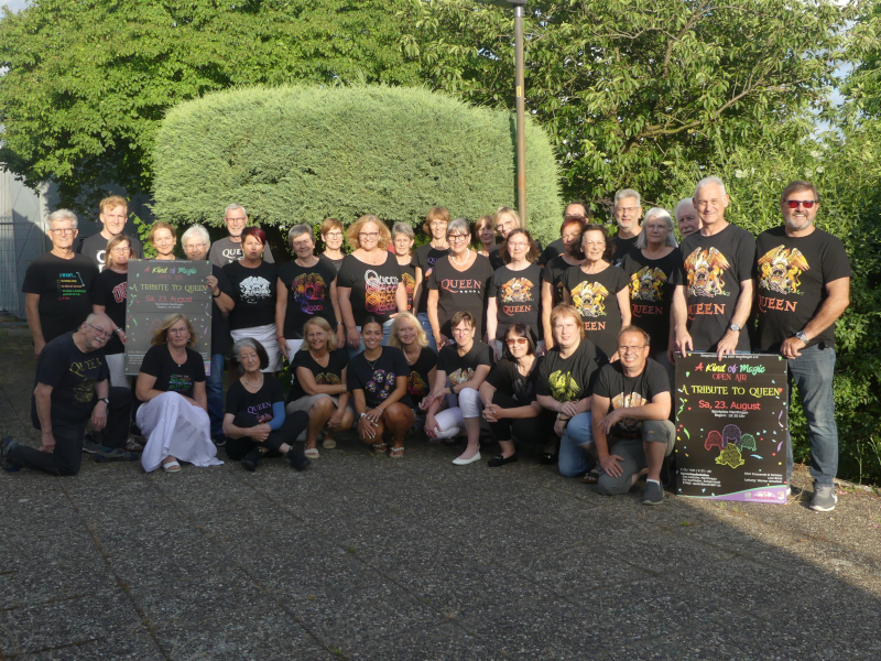 Der Chor Crescendo im Freien vor einer Hecke, viele in Queen-T-Shirts, daneben zwei Plakate zum Open-Air-Konzert